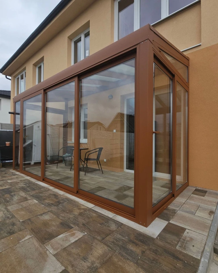 Eine moderne, verglaste Wintergartenkonstruktion mit braunem Rahmen, die an ein Einfamilienhaus angebaut ist.