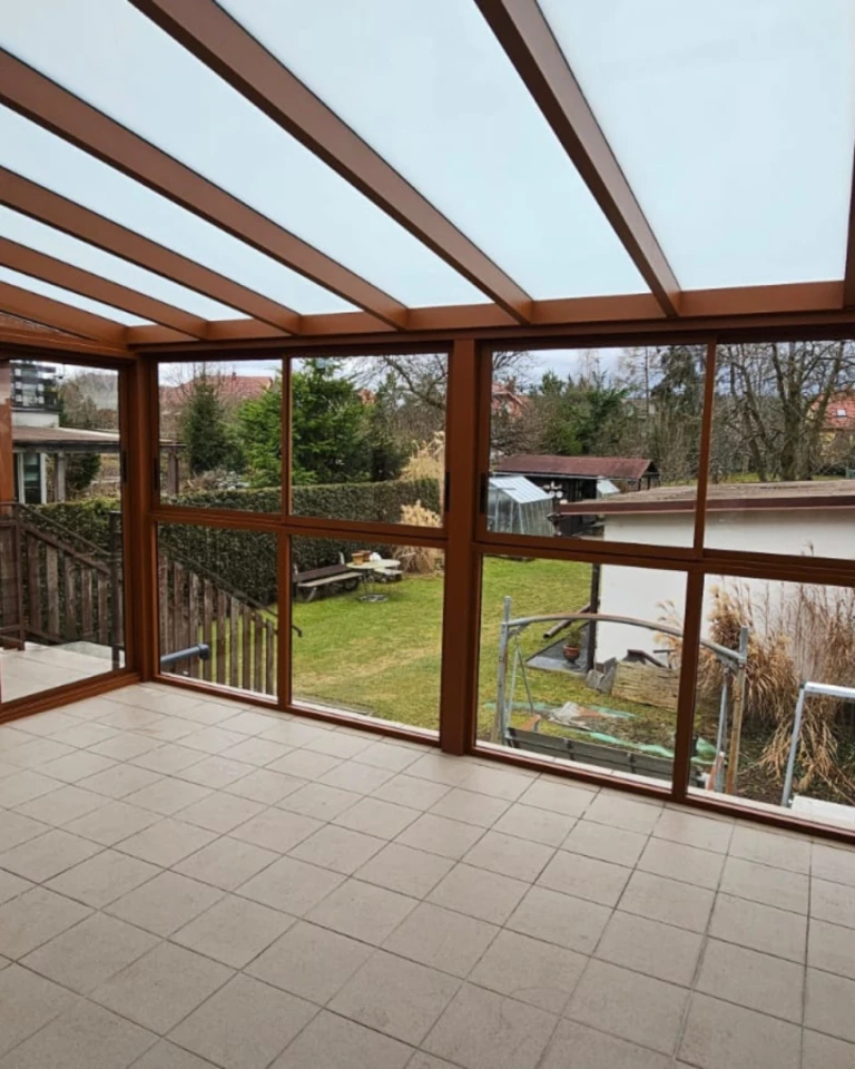 Blick aus dem Inneren einer braunen, verglasten Aluminium-Pergola mit Blick auf den Garten