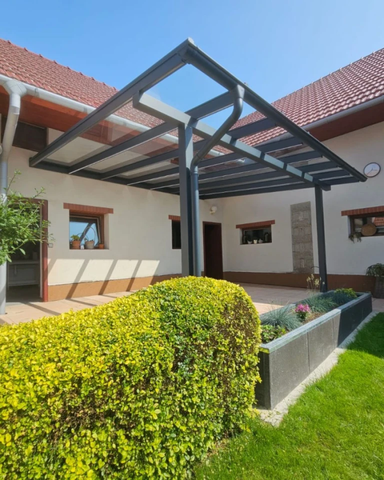 Eine moderne Aluminium-Pergola mit Glasdach, die an ein Einfamilienhaus mit gepflegtem Garten angebaut ist.