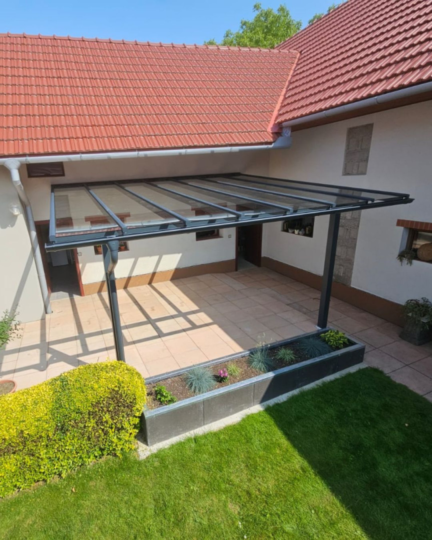 Eine moderne Aluminium-Pergola mit Glasdach, die an der Hauswand befestigt ist und über eine gepflasterte Terrasse verfügt.