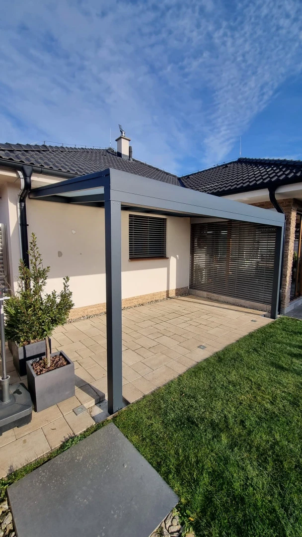Eine moderne anthrazitfarbene Aluminium-Pergola, die an das Haus angebaut ist und auf einer gepflasterten Terrasse mit Rasen steht.