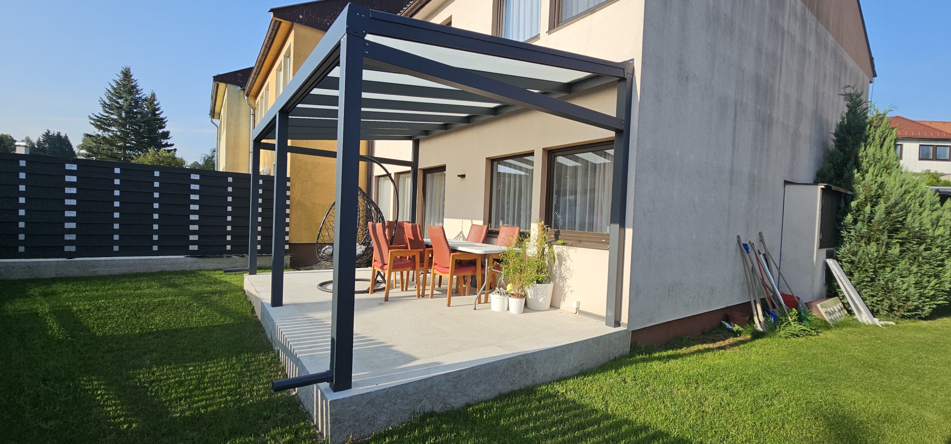 Eine moderne Aluminium-Pergola mit verglaster Überdachung auf der Betonterrasse eines Einfamilienhauses mit Gartenmöbeln.