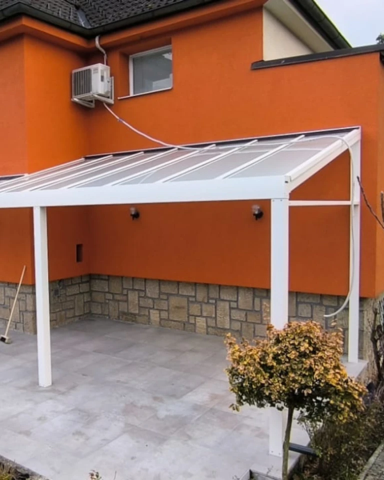Eine weiße Aluminium-Pergola mit Glasdach, die an ein orangefarbenes Haus angebaut ist