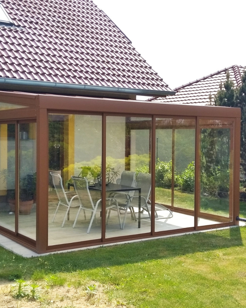 Ein verglaster Wintergarten aus Aluminium mit braunem Rahmen und Gartenmöbeln auf der Terrasse neben dem Haus