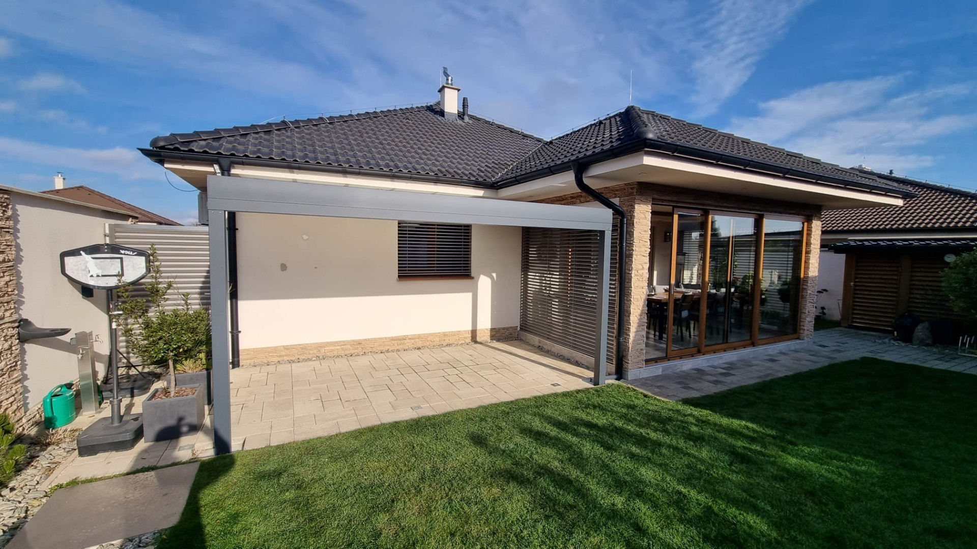 Eine moderne graue Aluminium-Pergola neben dem Bungalow auf der gepflasterten Terrasse am Gartenrand