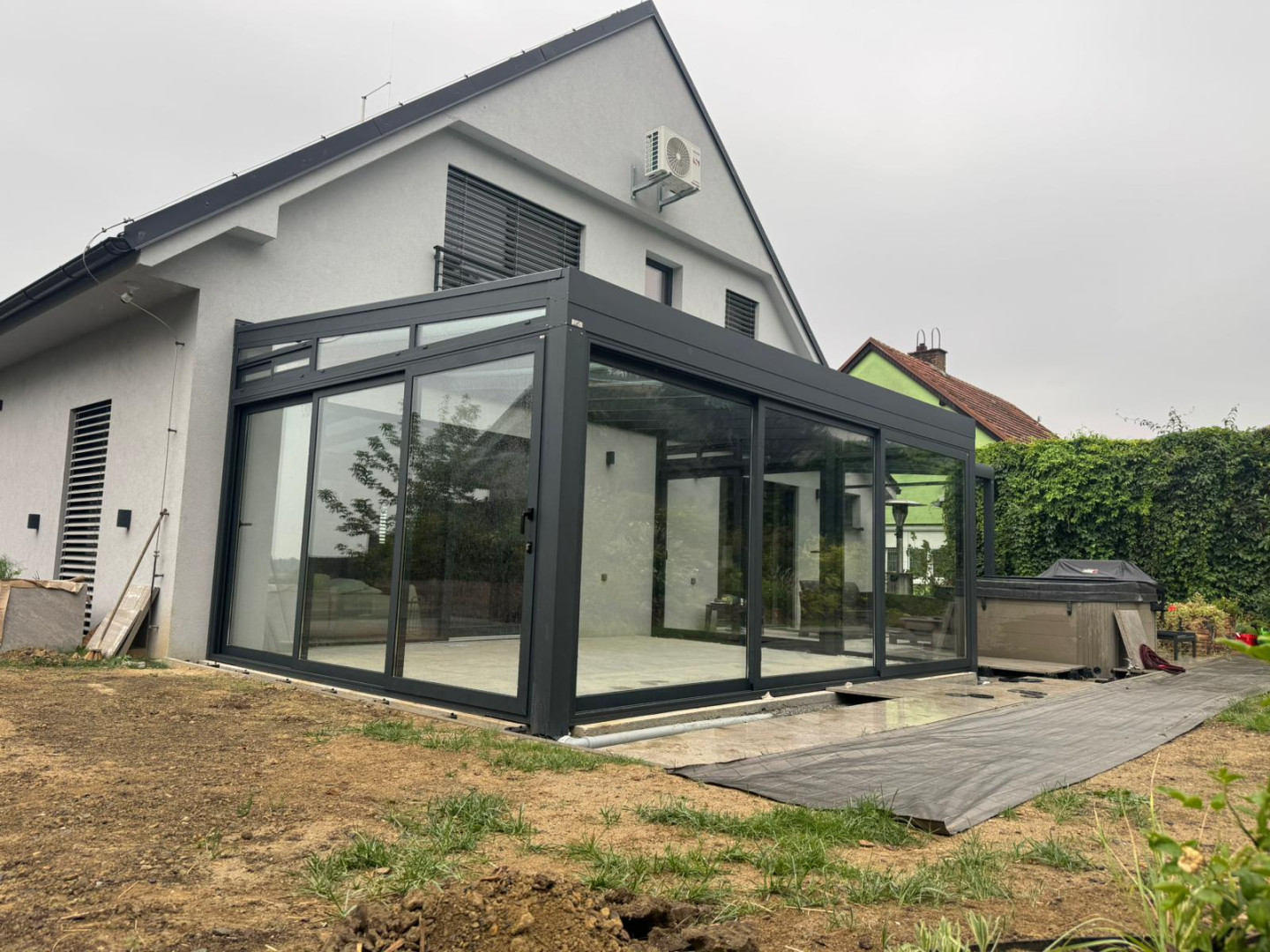 Eine moderne, verglaste Konstruktion, die an ein Einfamilienhaus angebaut ist, mit dunklem Rahmen und Schiebetüren.