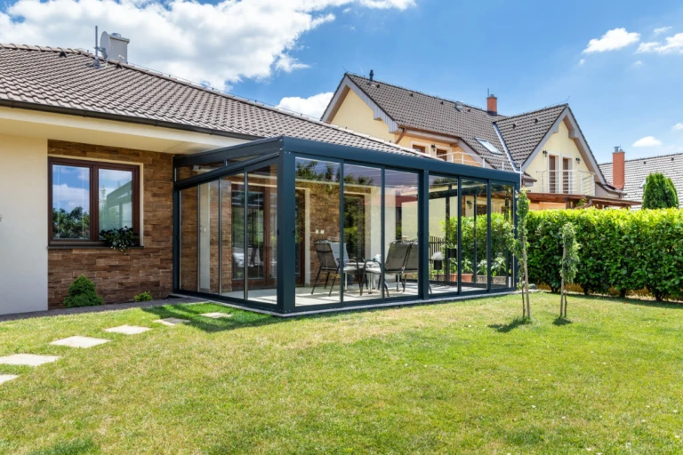 Ein moderner, verglaster Wintergarten mit dunkler Konstruktion, der an ein Einfamilienhaus angebaut ist und mit Gartenmöbeln ausgestattet ist.