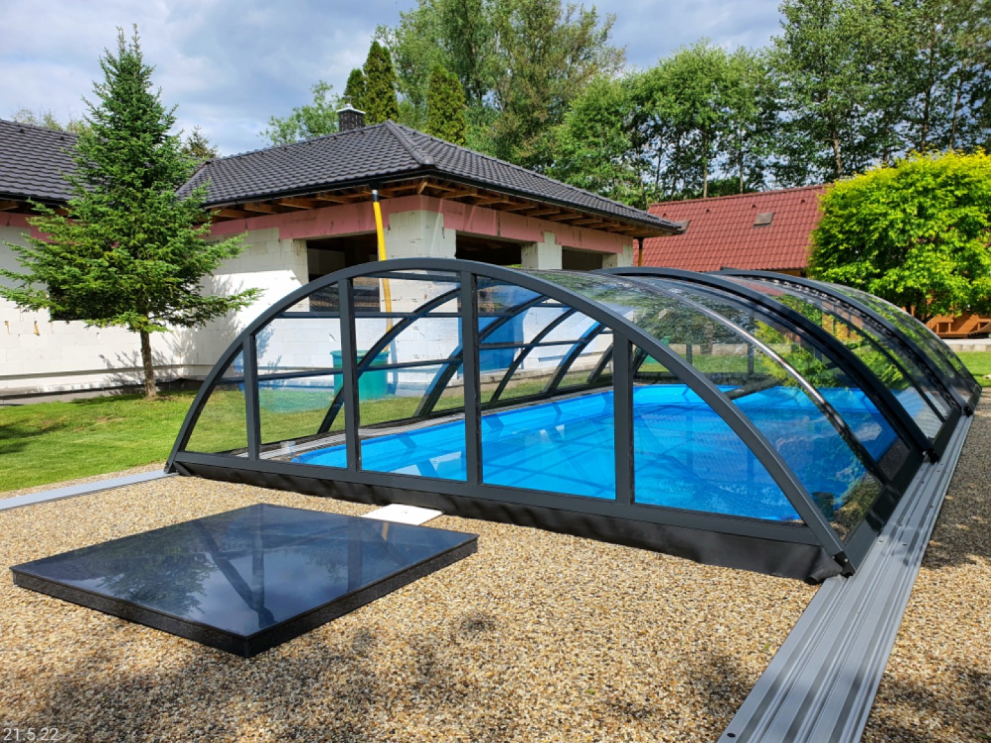 Anthrazitfarbene, bogenförmige Schwimmbadüberdachung mit Glasfüllung im Garten eines Einfamilienhauses.