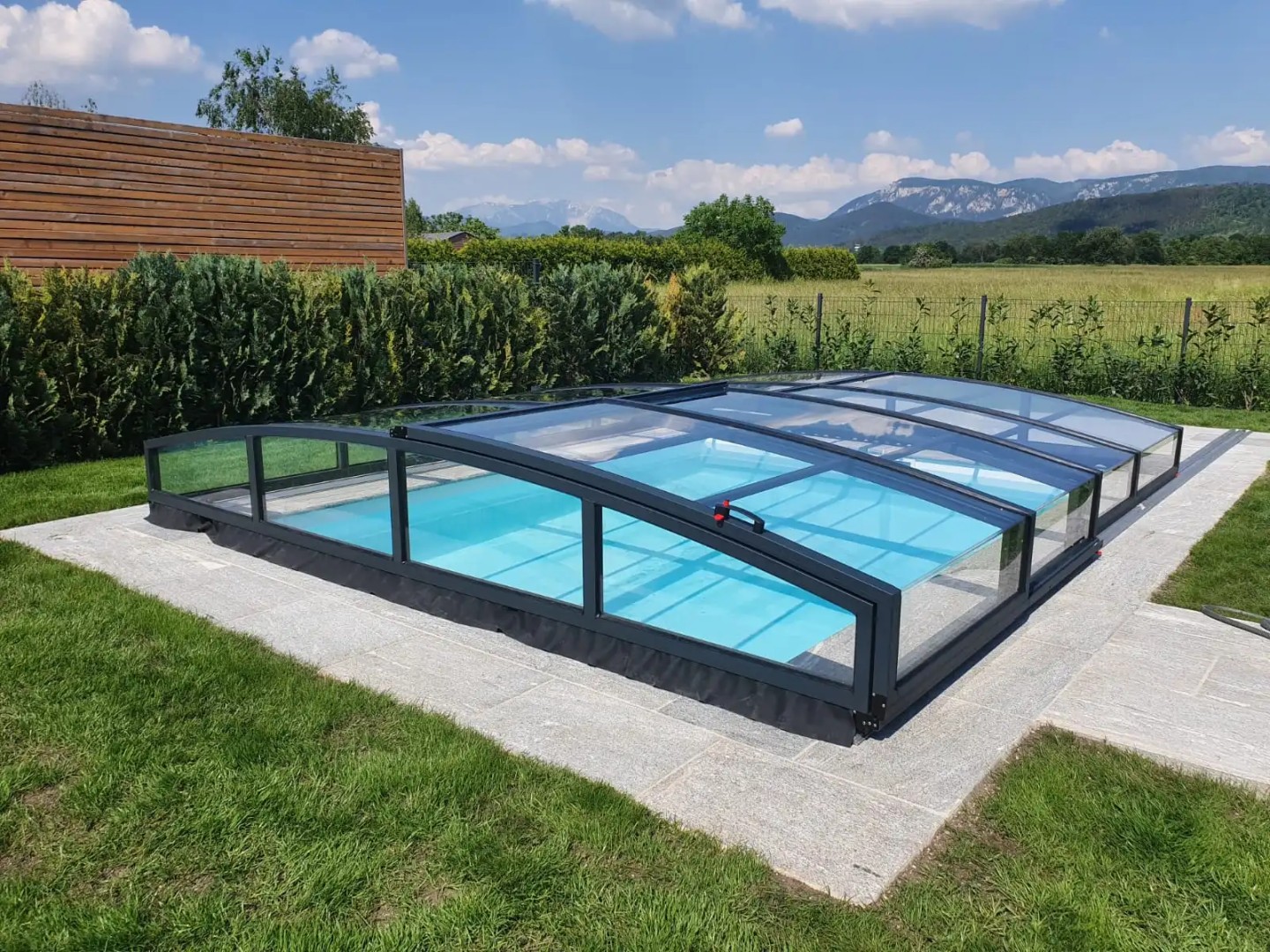 Niedrige anthrazitfarbene STAR-Schwimmbadüberdachung im Garten mit Blick auf die Berge