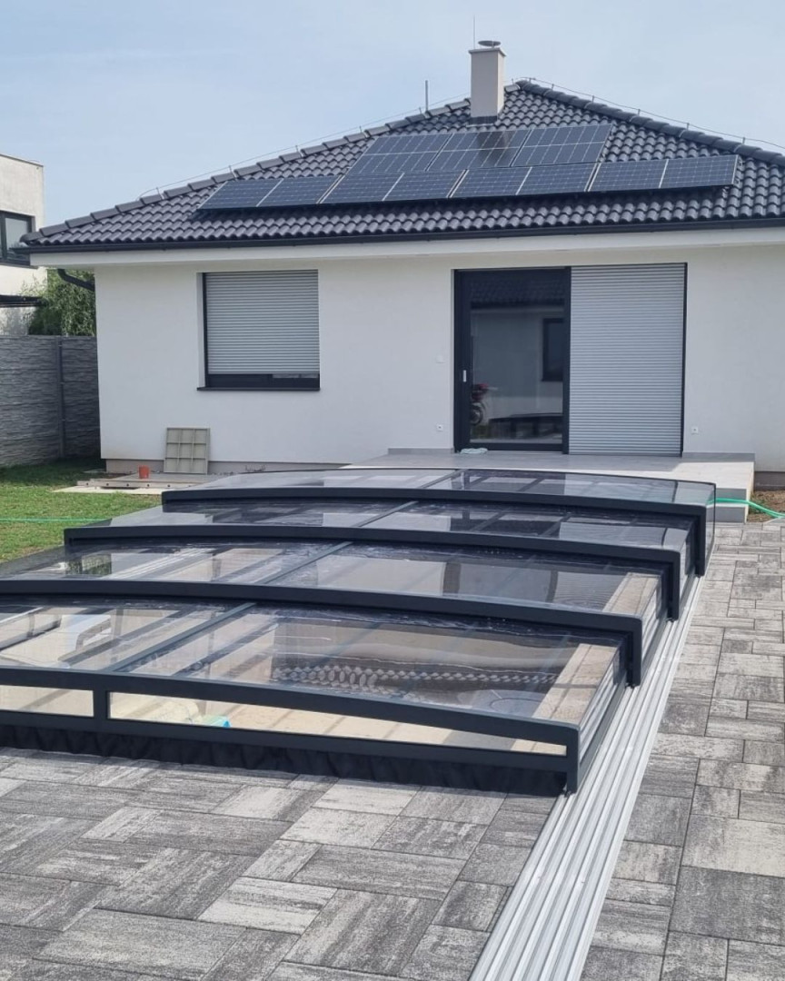 Eine moderne, niedrige Schwimmbadüberdachung in Anthrazit vor einem Einfamilienhaus mit Pflaster und Photovoltaikanlage.