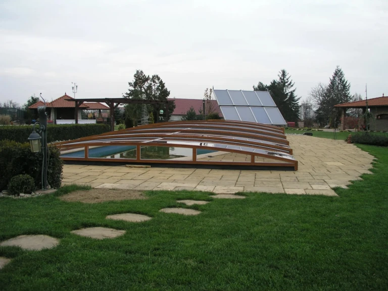 Eine niedrige Schwimmbadüberdachung aus Holz im Garten mit Steinpflaster und Sonnenkollektoren im Hintergrund.