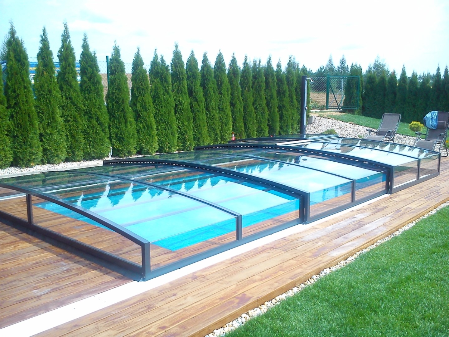 Eine moderne, niedrige Schwimmbadüberdachung mit transparenter Verkleidung auf einer von Grün umgebenen Holzterrasse