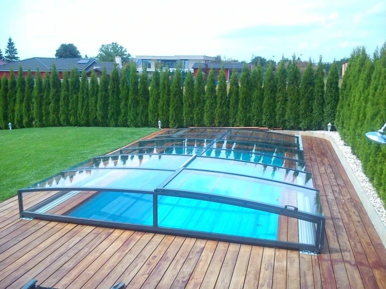 Eine moderne, niedrige Schwimmbadüberdachung mit transparenter Verkleidung auf einer Holzterrasse im Garten mit einer Hecke.