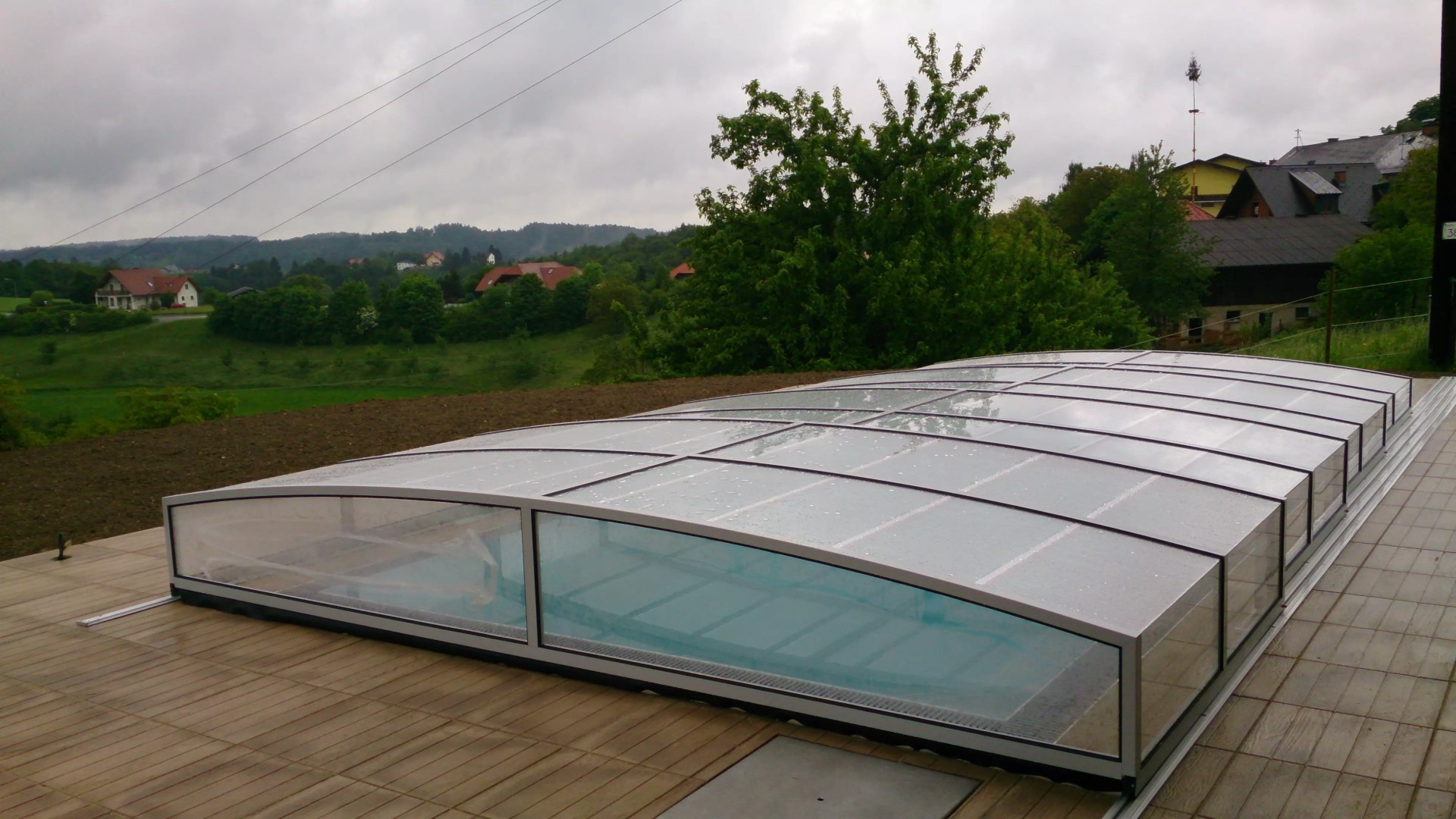 Eine moderne, niedrige STAR-Schwimmbadüberdachung auf der Terrasse mit Blick auf die hügelige Landschaft und die Häuser.