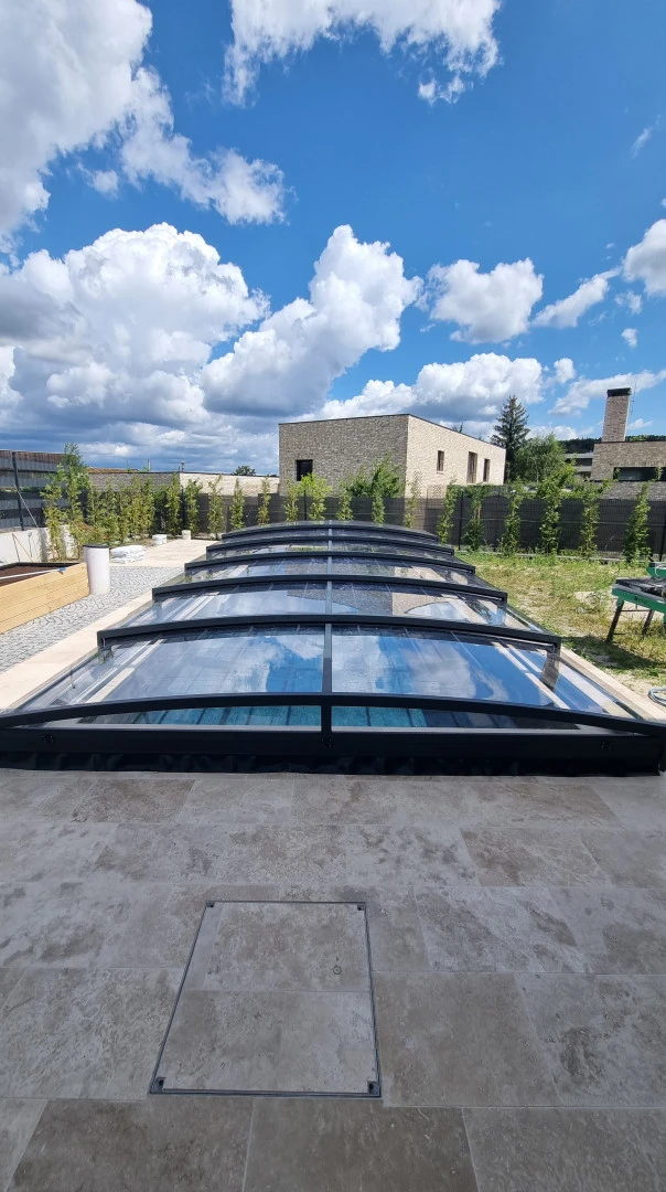 Eine moderne, niedrige Schwimmbadüberdachung mit schwarzer Konstruktion und transparenter Verkleidung im Garten eines Einfamilienhauses.