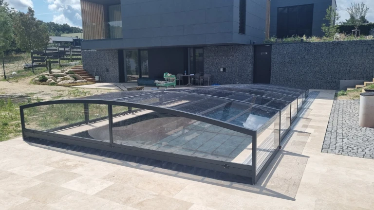 Eine moderne, niedrige Schwimmbadüberdachung in dunkler Farbe vor einem modernen Haus mit Steinverkleidung.