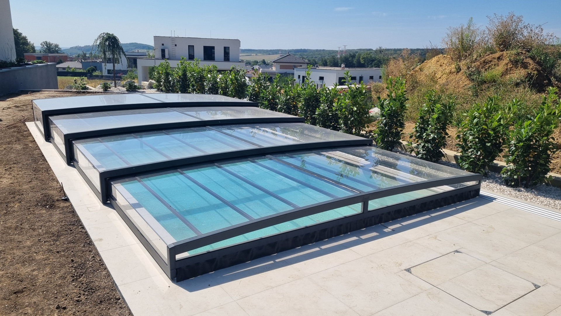 Moderne, niedrige Teleskop-Schwimmbadüberdachung mit anthrazitfarbener Konstruktion und transparenter Verkleidung auf der Terrasse eines Einfamilienhauses.
