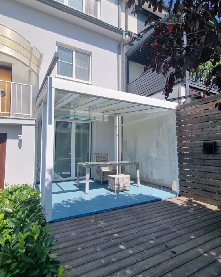 Eine weiße, moderne Aluminium-Pergola mit Glasdach auf der Terrasse eines Einfamilienhauses mit Gartenmöbeln.