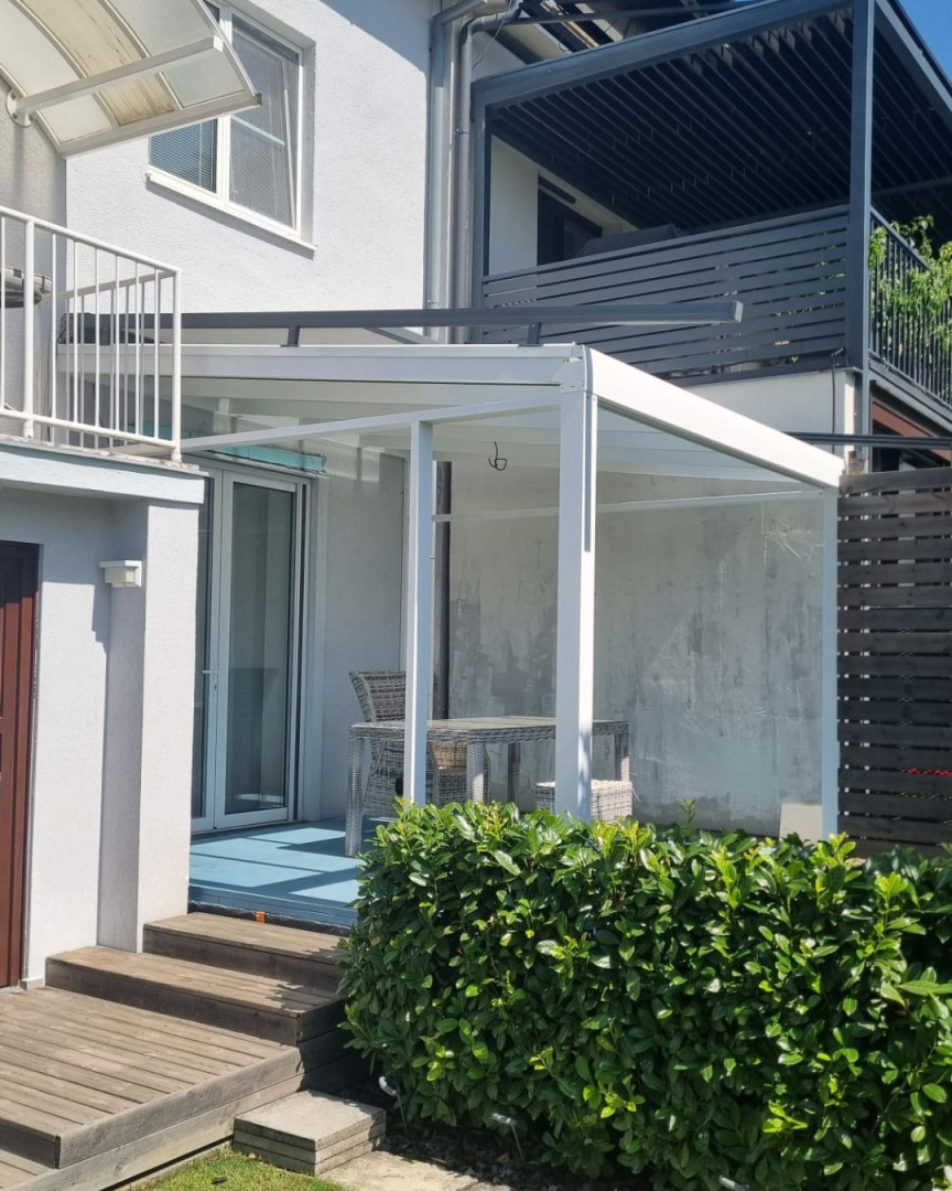 Eine weiße, moderne Aluminium-Pergola mit Glasdach an einem Einfamilienhaus mit Terrassenmöbeln.