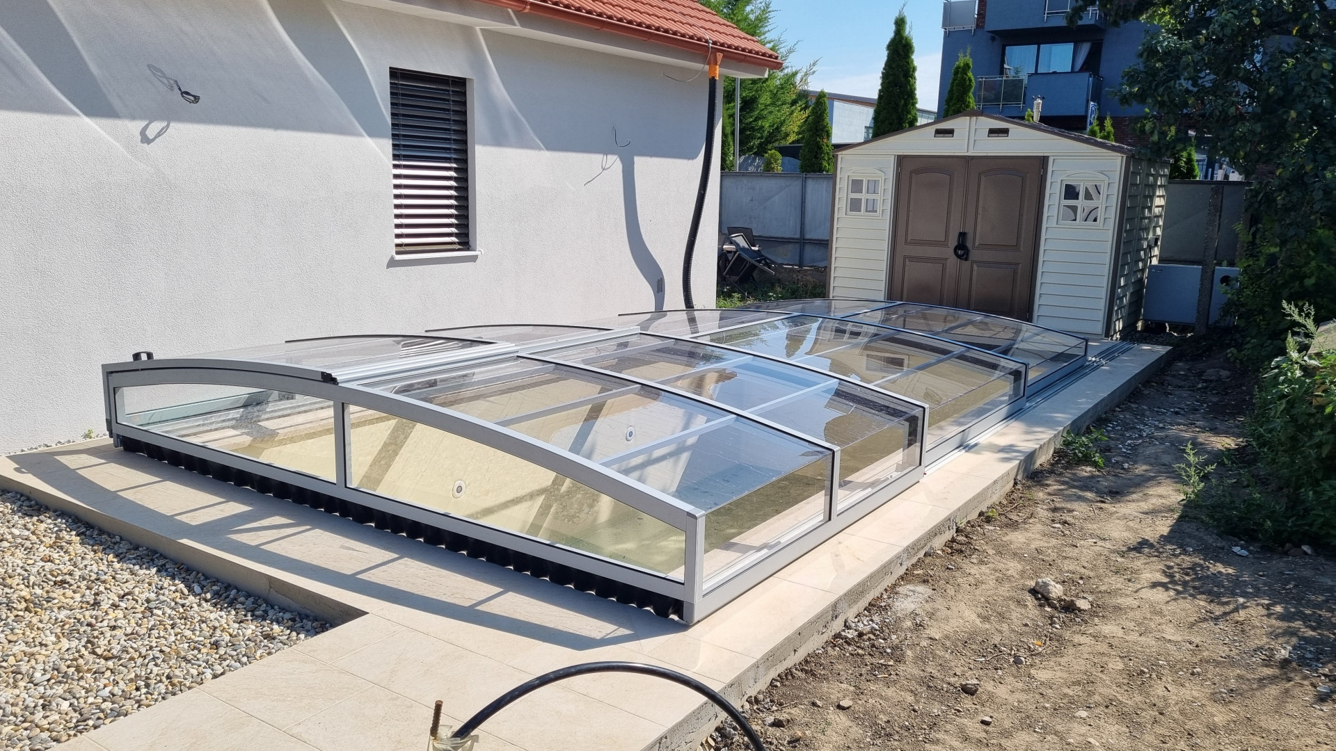 Eine niedrige Segmentüberdachung für ein Schwimmbecken mit silberner Konstruktion und transparentem Polycarbonat bei einem Einfamilienhaus in Senec.