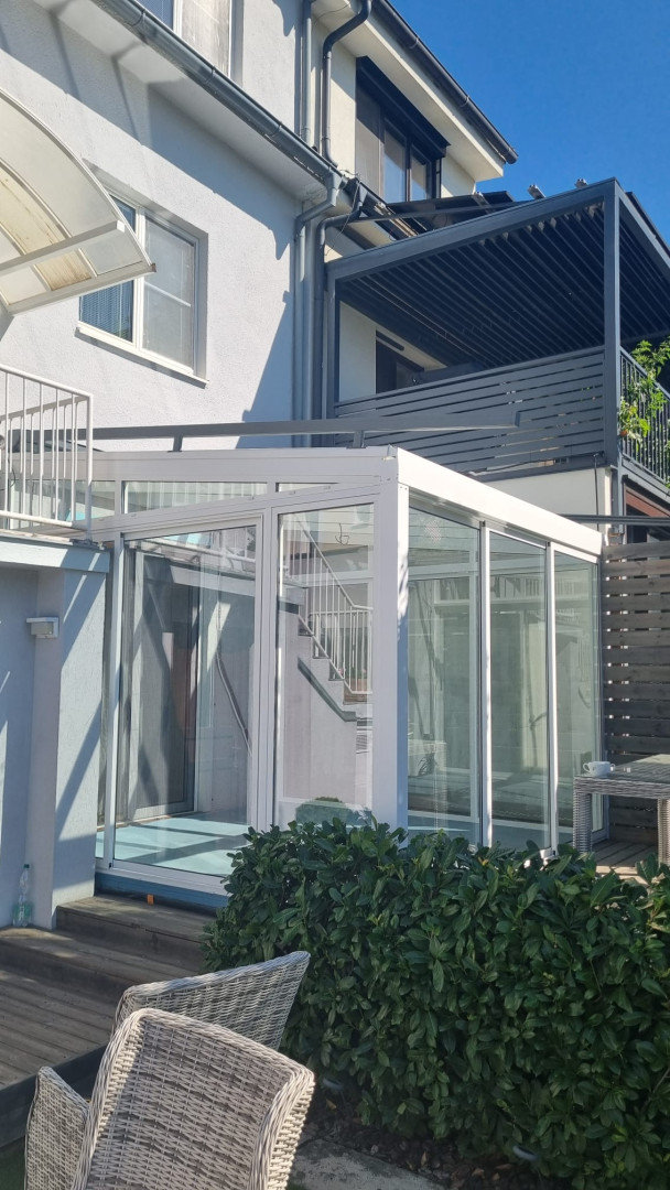 Ein moderner, weißer, verglaster Wintergarten, der an ein Einfamilienhaus angebaut ist und einen Blick auf die Terrasse und die grüne Umgebung bietet.
