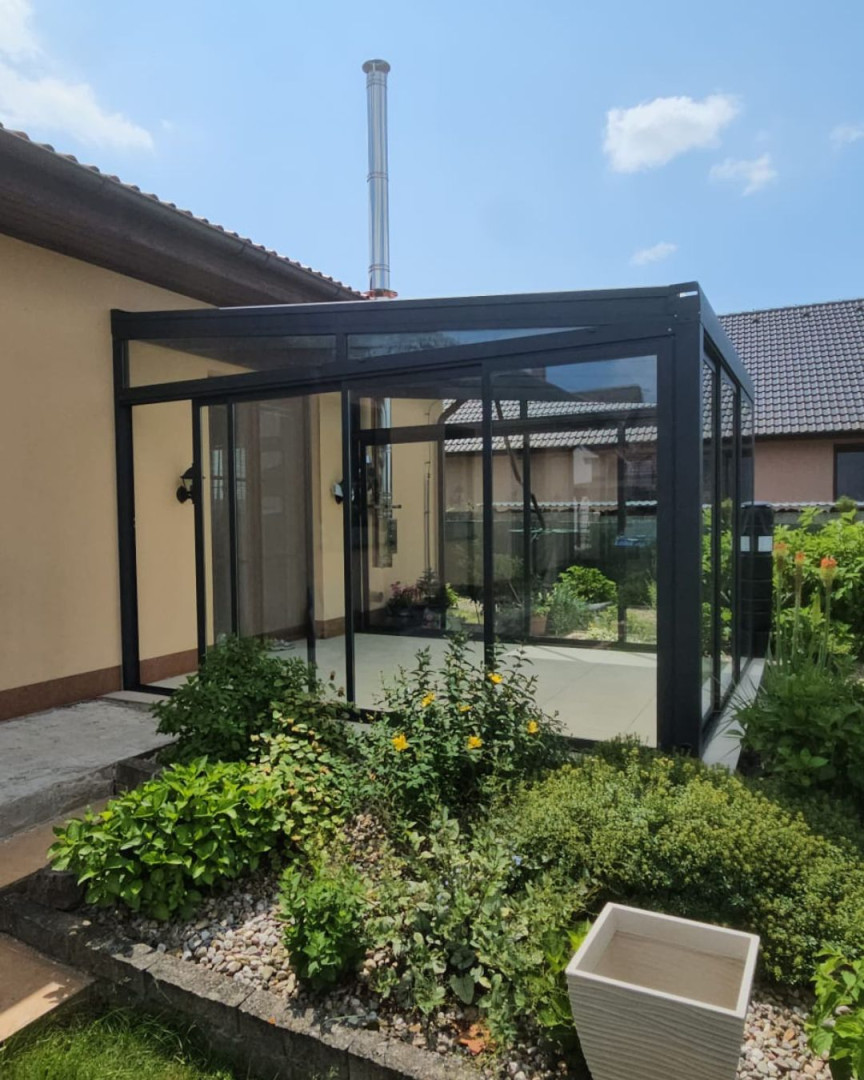 Ein moderner, verglaster Anbau an ein Einfamilienhaus mit schwarzem Rahmen und Schiebetüren, umgeben von einem Garten.