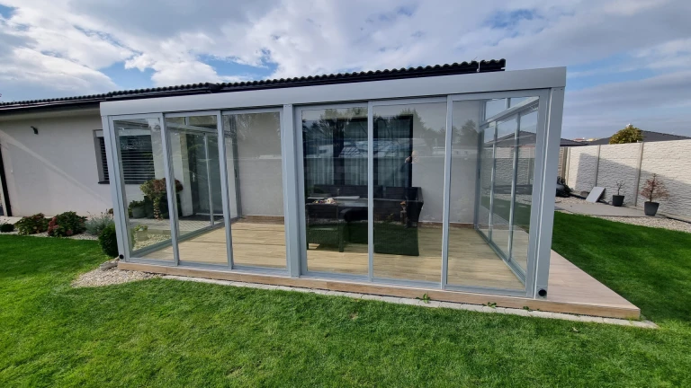 Ein moderner, verglannter Wintergarten mit grauer Konstruktion auf einer Holzterrasse neben einem Einfamilienhaus