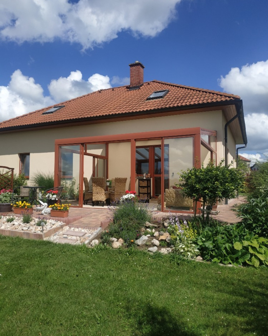 Ein moderner, verglaster Wintergarten mit brauner Konstruktion, der an ein Einfamilienhaus mit gepflegtem Garten angebaut ist.