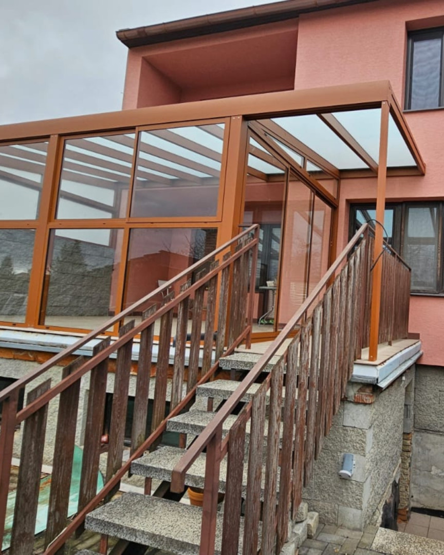 Eine verglaste, braune Aluminium-Pergola, die auf der Terrasse eines Hauses mit Treppe montiert ist