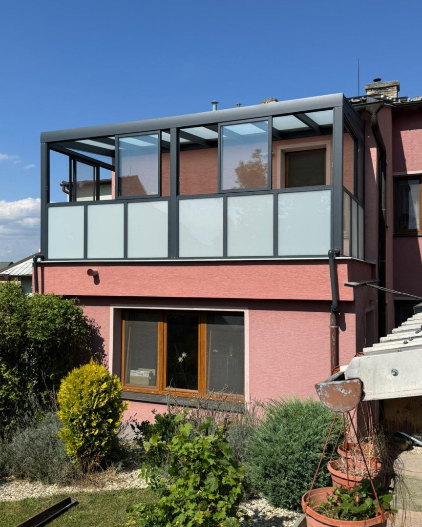 Eine moderne, verglaste Pergola mit dunkelgrauem Aluminiumrahmen und Milchglas auf dem Balkon eines Einfamilienhauses.