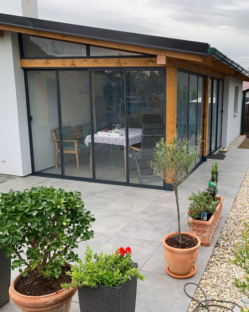 Moderne Terrassenverglasung mit Schiebesystem und Holzkonstruktion an einem Einfamilienhaus, im Vordergrund stehen Blumentöpfe.