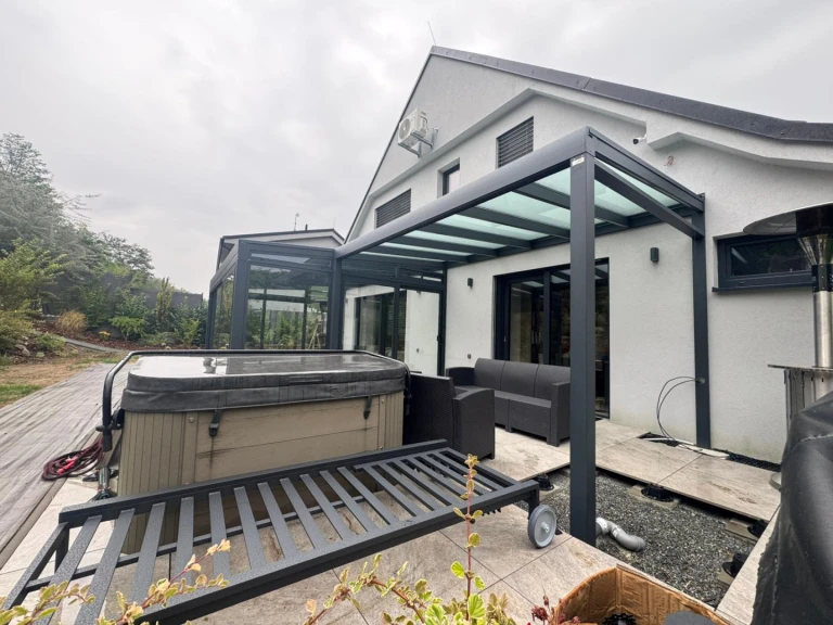 Eine moderne, verglaste Pergola und ein Wintergarten an einem Einfamilienhaus mit Whirlpool und Rattanmöbeln auf der Terrasse.