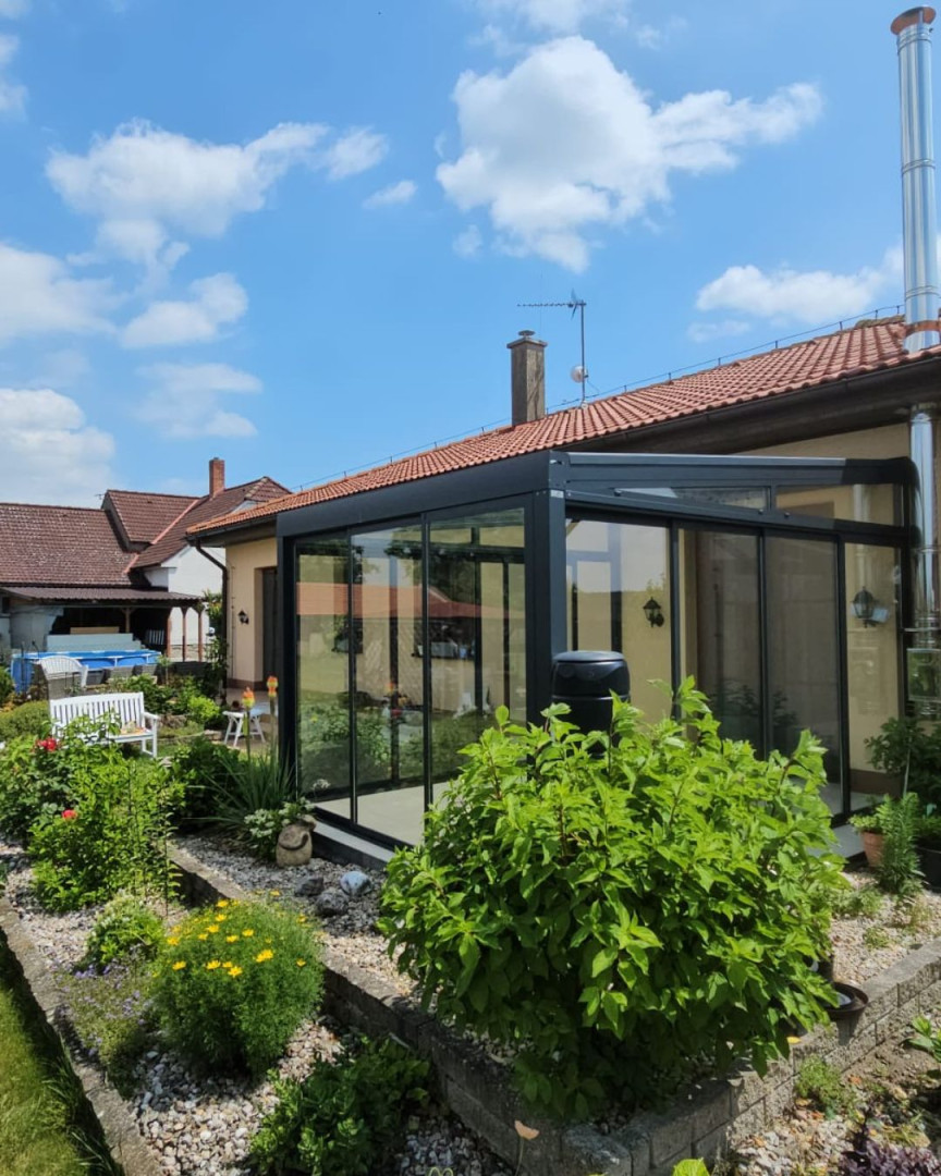 Ein moderner, verglaster Wintergarten mit anthrazitfarbener Konstruktion an einem Einfamilienhaus mit gepflegtem Garten